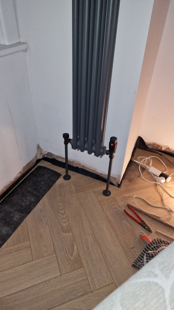 A room under construction, featuring a gray radiator against a white wall with electrical floor tapping points near the fl...