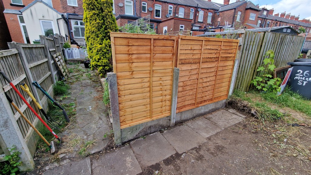 An unfinished fence at the end of a dirt path in an urban back garden, surrounded by wooden fences and rubbish bins, with ...