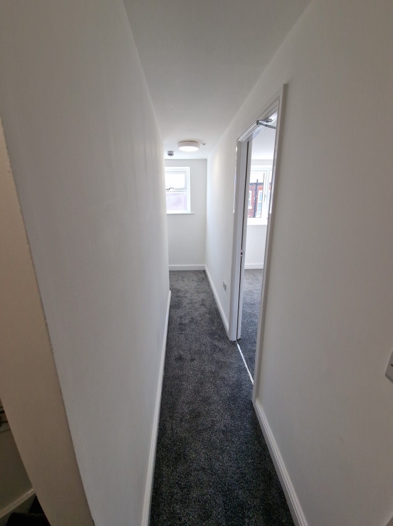 The image depicts a narrow, white hallway with gray carpeting and a door at the end, which is slightly ajar, revealing a l...