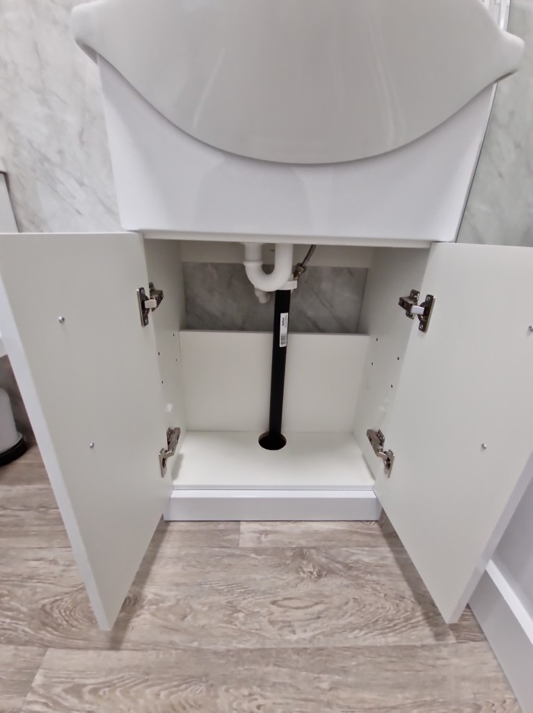 The alt text for this image is "A bathroom cabinet with furniture-style doors, open, revealing the plumbing in the space i...