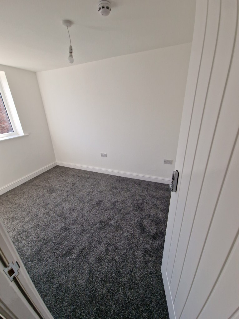 A room with a window, white walls and ceiling, and gray carpet. There are two light bulbs hanging from the ceiling, and a ...