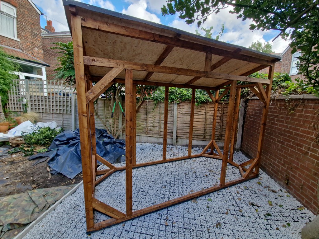 The wooden structure, likely a gazebo or small garden shelter, is currently undergoing construction. It is supported by wo...